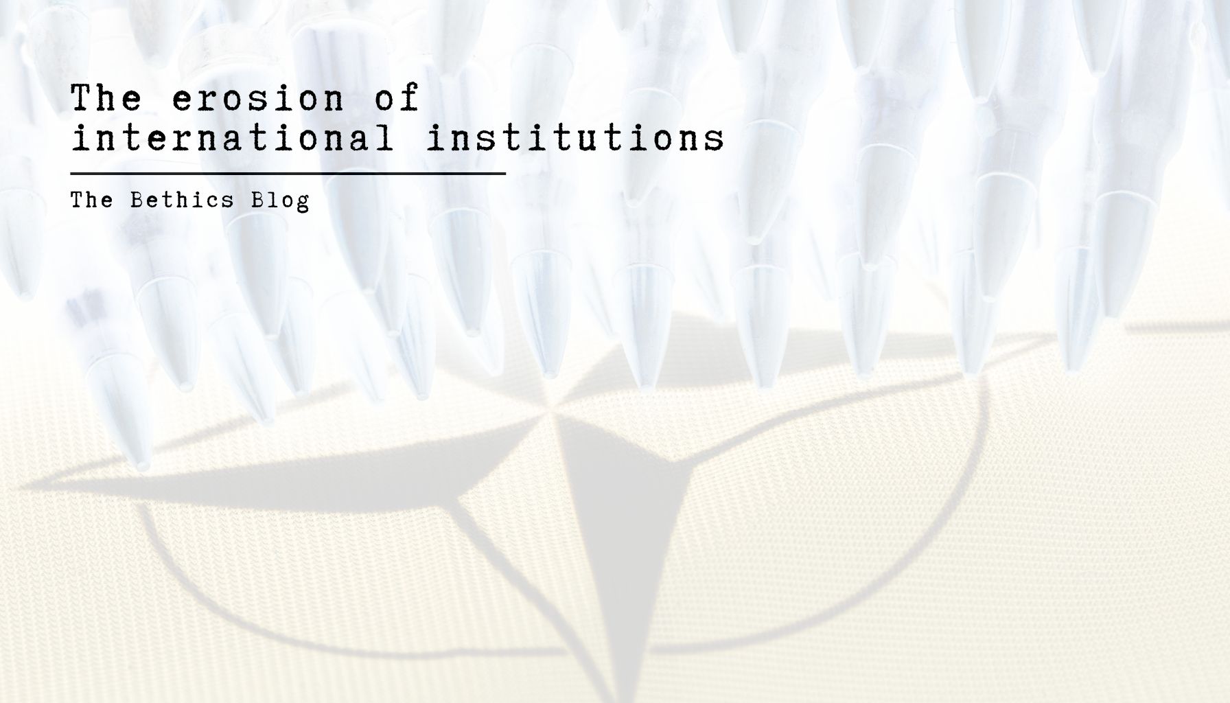 A high-key, minimalist image featuring a row of sharp, bullet-like white shapes pointing downward toward a faded, translucent NATO compass rose. The text "The erosion of international institutions" is written in a black typewriter font in the upper left corner, underscored by the words "The Bethics Blog."